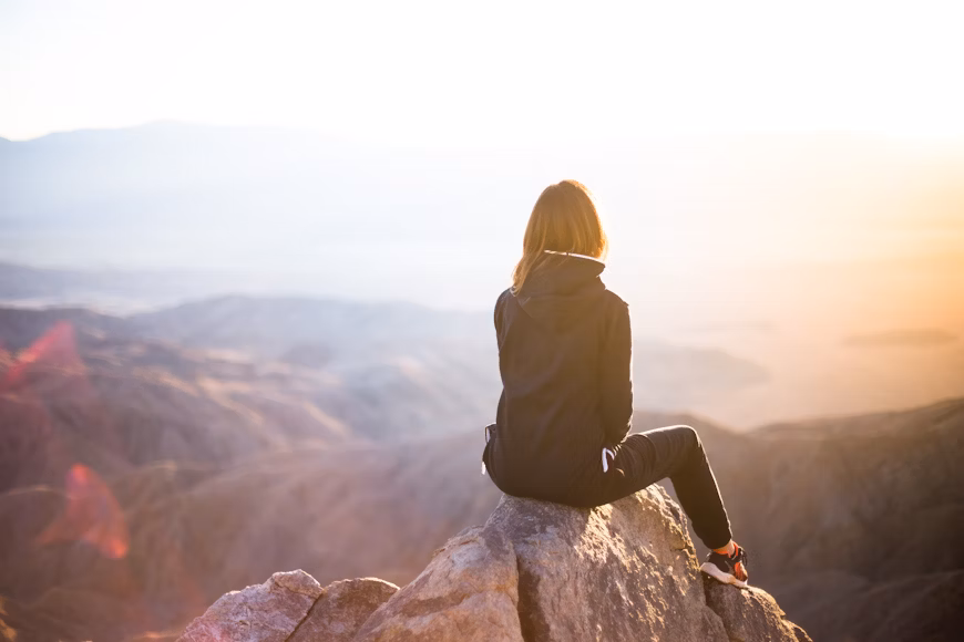 Young woman sitting on top of mountain, looking out over a beautiful horizon contemplating her future goals with a growth mindset