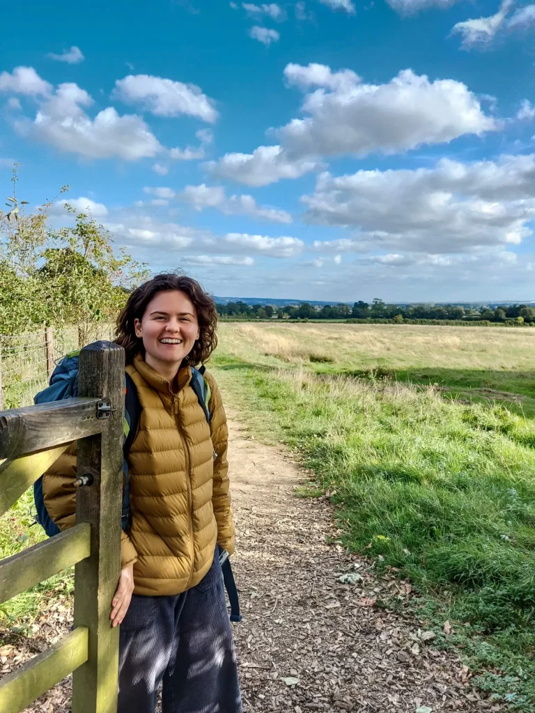 Lauren smiling outdoors in a field, holding a gate open for a friend on a sunny summer's walk